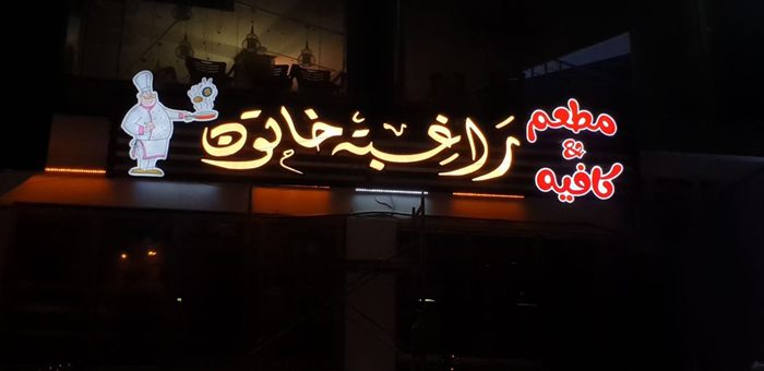 لوحة مضيئة ليلاً بحروف عربية صفراء وحمراء مكتوب عليها راغبة خاتون مطعم وكافيه