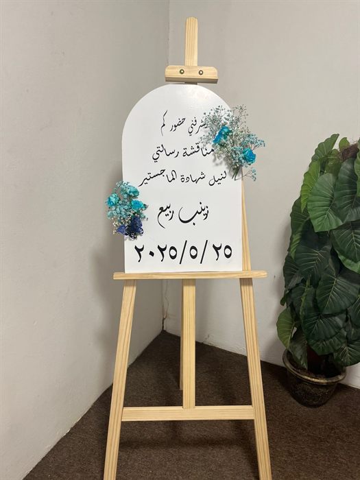 لوحة بيضاء مقوسة عليها دعوة لمناقشة رسالة ماجستير مع زينة ورود زرقاء