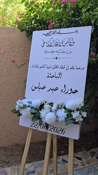 لوحة بيضاء على حامل خشبي عليها ترحيب باسم الباحثة وتاريخ المناقشة